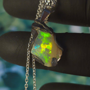 Raw uncut Ethiopian Welo opal pendant. Colour: blue, red, with green and yellow accents, vivid. Body: white. Held in a gloved hand against a green background.