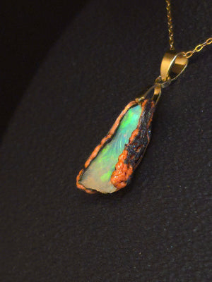 Raw uncut Ethiopian Welo opal pendant. Colour: green leading, red cutting in, blue at the edges, strong. Body: crystal clear. Lying on a dark surface.