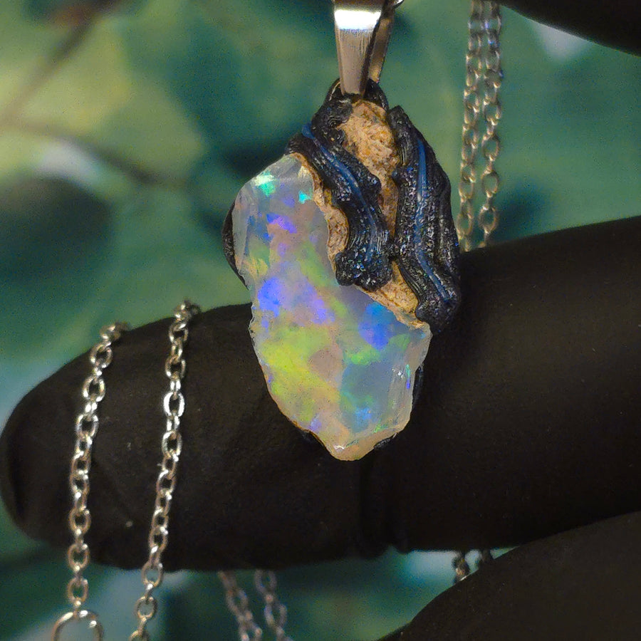 Raw uncut Ethiopian Welo opal pendant. Colour: blue, teal, with green hints, moderate. Body: light blue. Held in a gloved hand against a green background.